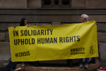 Kaksi mielenosoittajaa pitää Amnestyn banneria käsissään, teksti: In Solidarity Uphold Human Rights