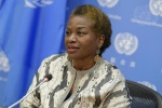Nainen puolilähikuvassa, taustalla YK:n logoja sinisellä pohjalla.