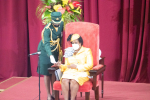Barbadoksen kenraalikuvernööri Sandra Mason istuu keltaisessa asussa juhlavassa tuolissa, univormuasuinen nainen ojentaa jotain hänen käteensä.