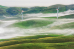 Kaksi tuulivoimalaa sumuisten vihreiden kukkuloiden keskellä.