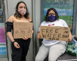 Kaksi kasvosuojaimin suojautunutta mielenosoittajaa, joilla "Free water" -tekstillä varustetut kyltit.
