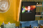 Mies puolilähikuvassa seinällä olevalla näyttöruudulla, vieressä YK:n logo.