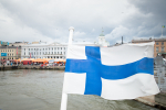 Suomen lippu, taustalla merta ja rakennuksia.