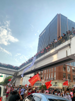 Mielenosoittajia punaisten lippujen kera kadulla, yläpuolella sillalla lisää ihmisiä.
