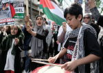 Poika palestiinalaishuivissa rummuttaa keskellä mielenosoitusta. Taustalla ihmiset liehuttavat Palestiinan lippua.