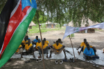 Joukko tummaihoisia poikia tai nuoria miehiä istuu puiden varjossa, etualalla näkyy osa Etelä-Sudanin lipusta.