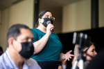Maskilla suojautunut nainen puhuu mikrofoniin yleisön keskellä.